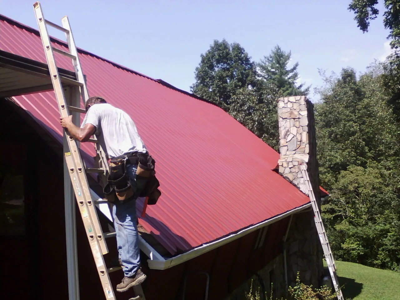 Roofer on a ladder beside a red metal roof and stone chimney — Soffit & Fascia Repair by Roofing Brothers in Hopewell Junction